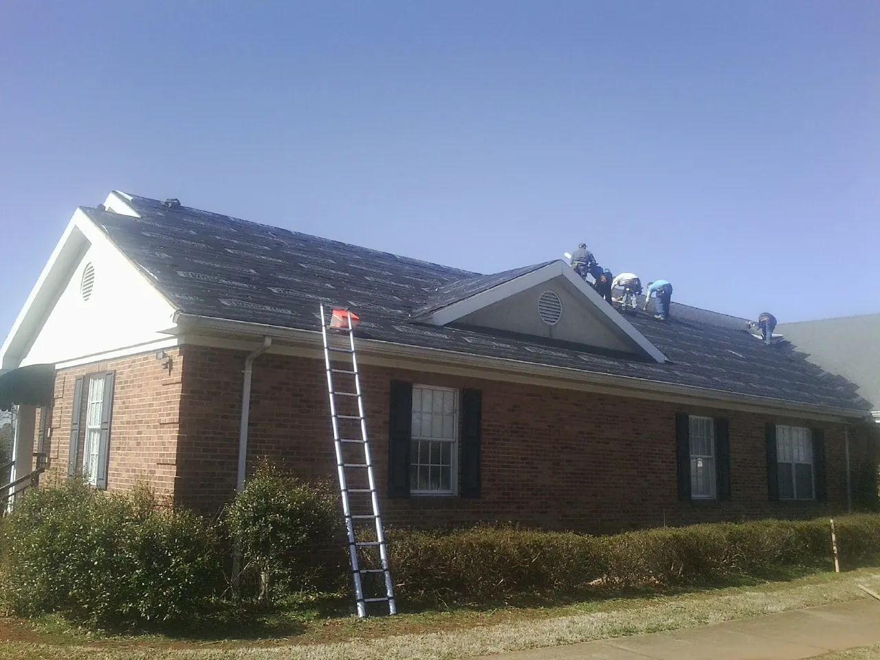 Crew tearing shingles off a brick ranch roof with a ladder set — Metal Roof Repair work by our crew in Avera