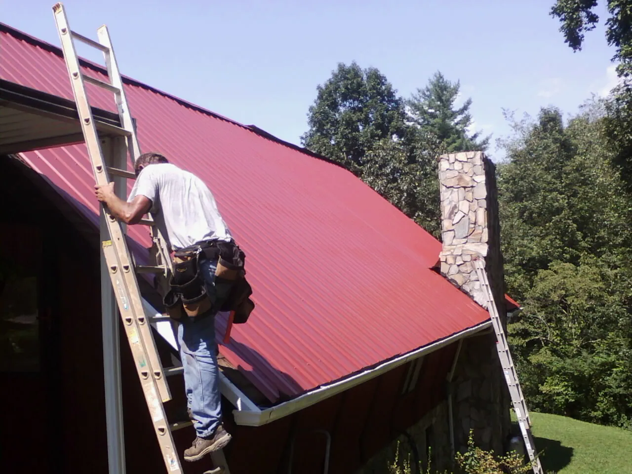 Roofer on a ladder beside a red metal roof and stone chimney — Soffit & Fascia Repair by Roofing Brothers in Avera